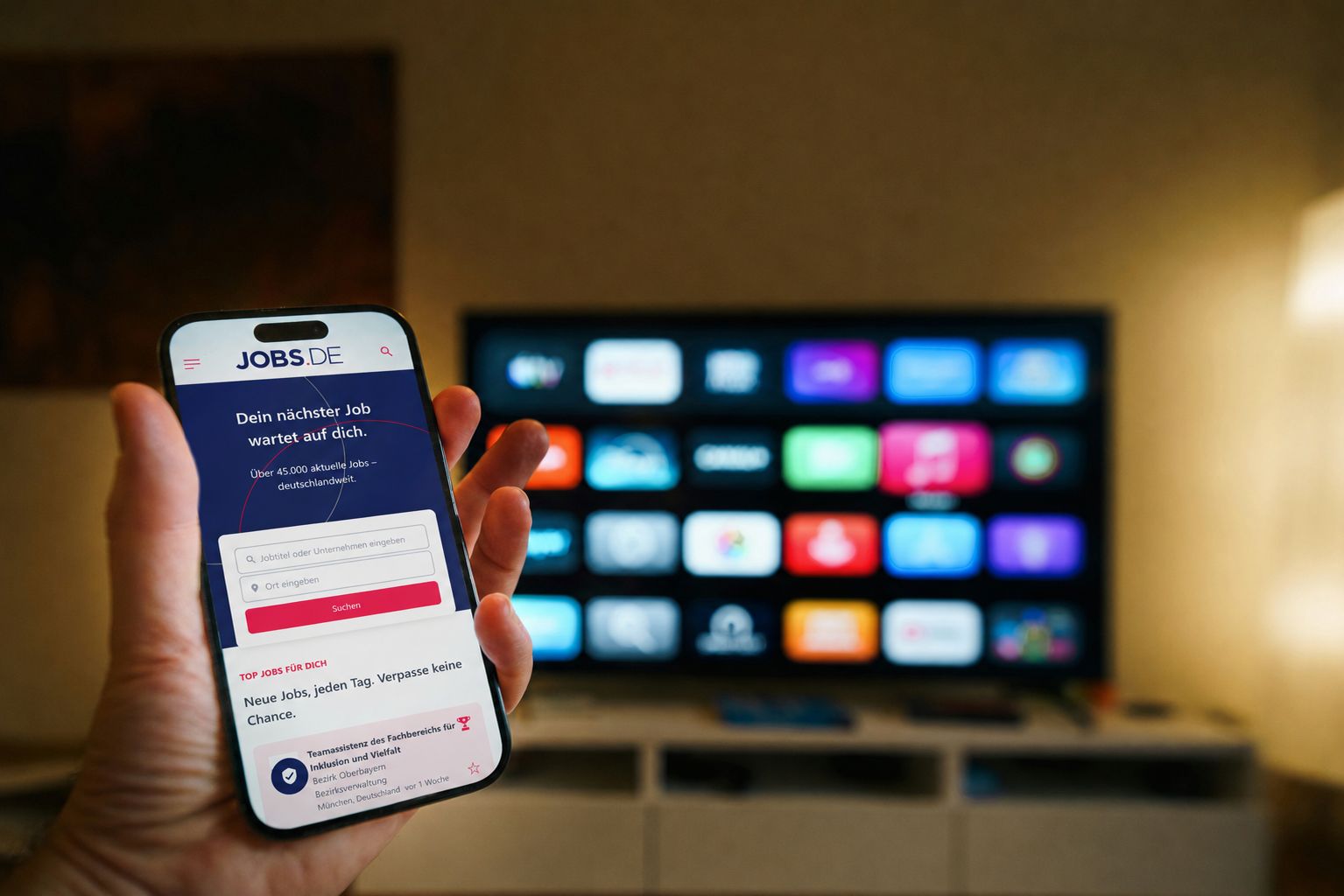Eine Hand hält ein Smartphone mit der JOBS.DE App, im Hintergrund ein Bildschirm mit digitalen Medienangeboten